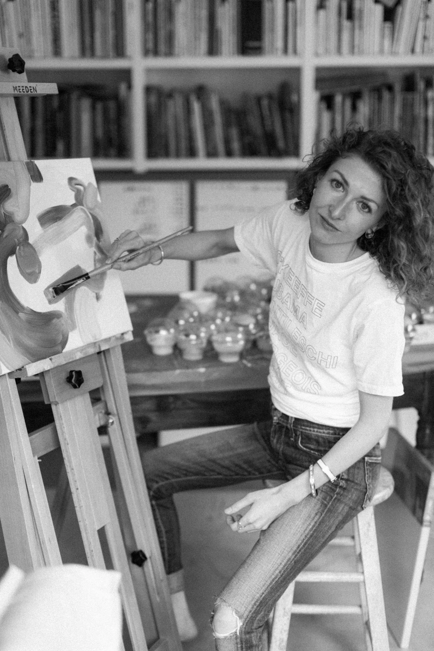 A black-and-white portrait of Molly Dimeo seated at her easel in the studio.