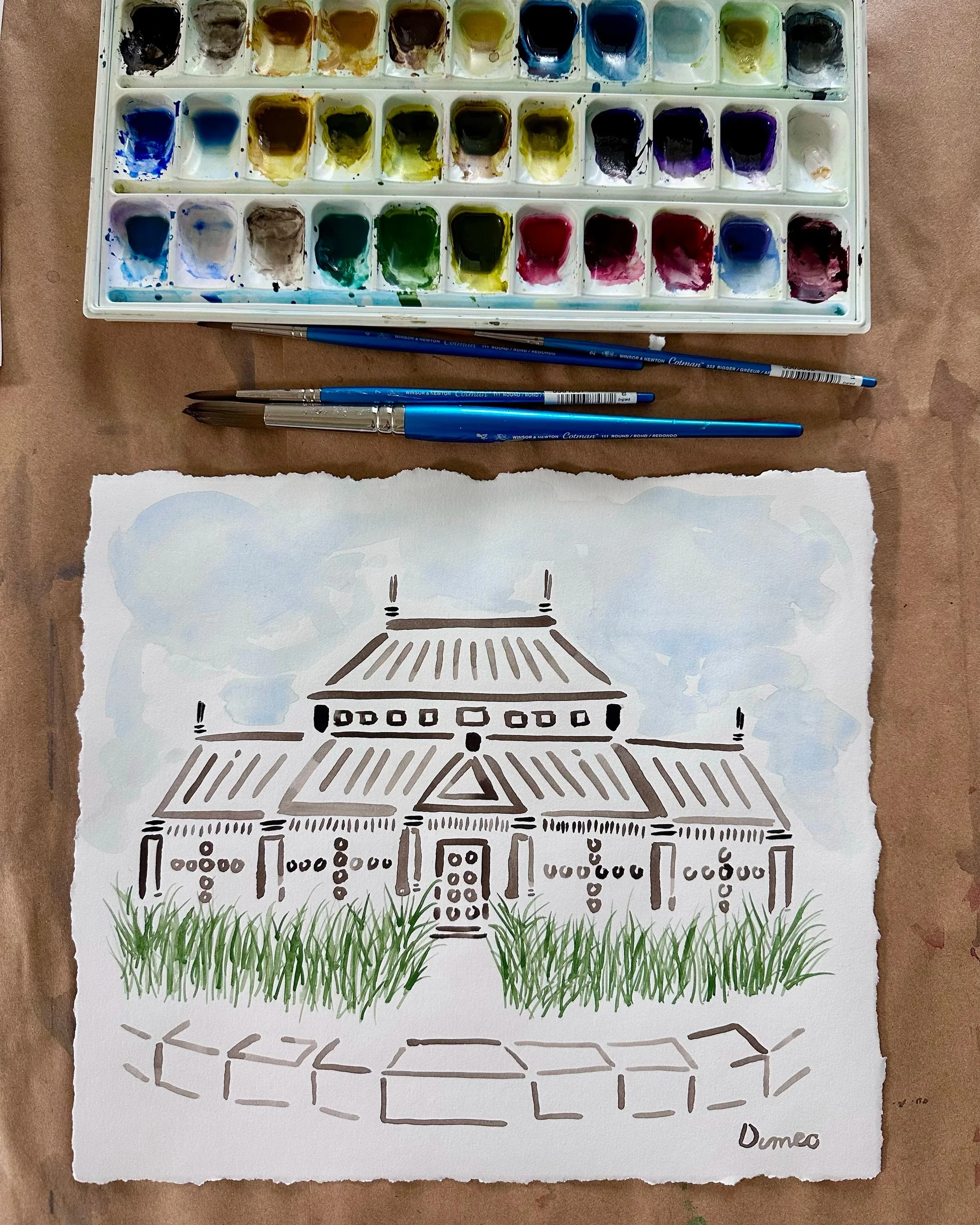 A watercolor painting of a building on deckled paper, with paintbrushes and a watercolor palette.